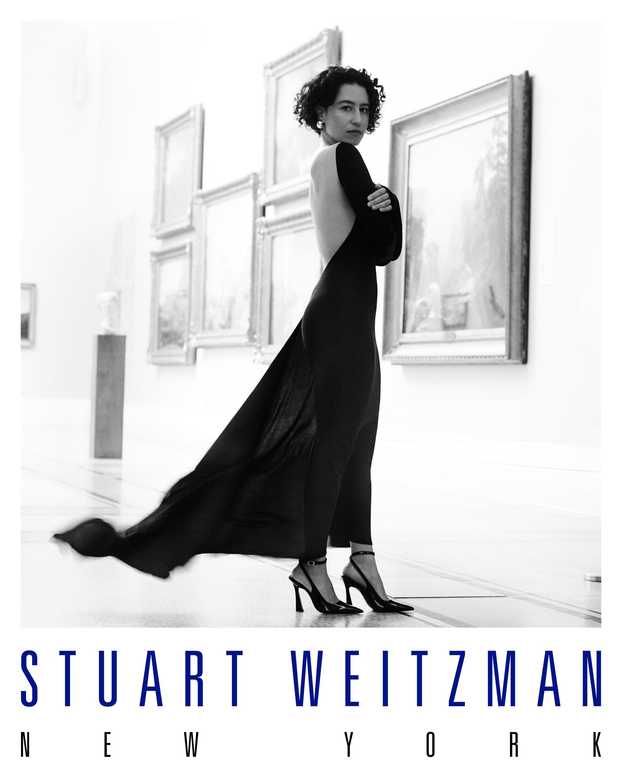 Ilana Glazer in a long black backless dress and heels posing inside a gallery for Stuart Weitzman Fall 2025 ad campaign