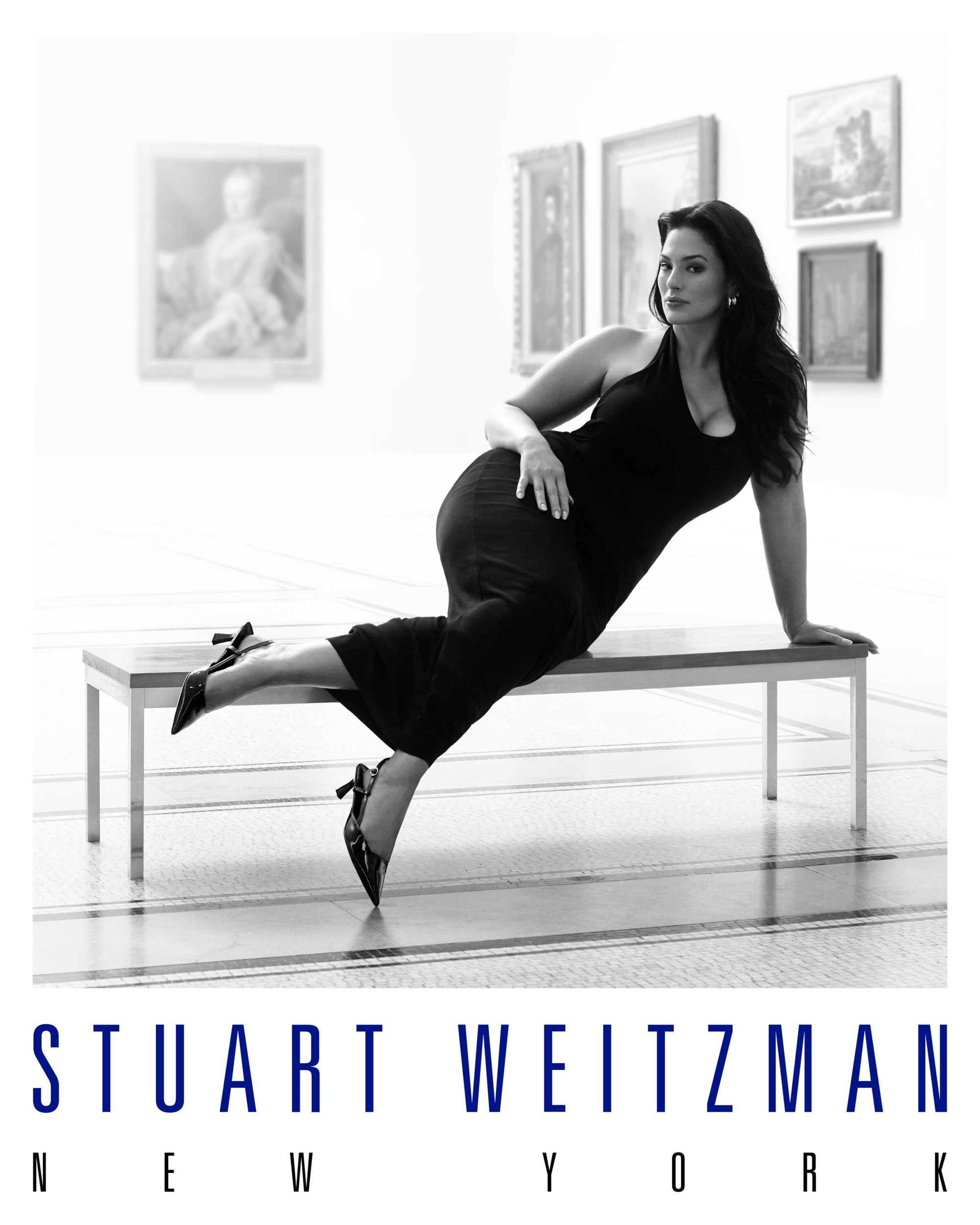Ashley Graham reclining on a gallery bench wearing a dark dress and heels in a black and white campaign image for Stuart Weitzman.