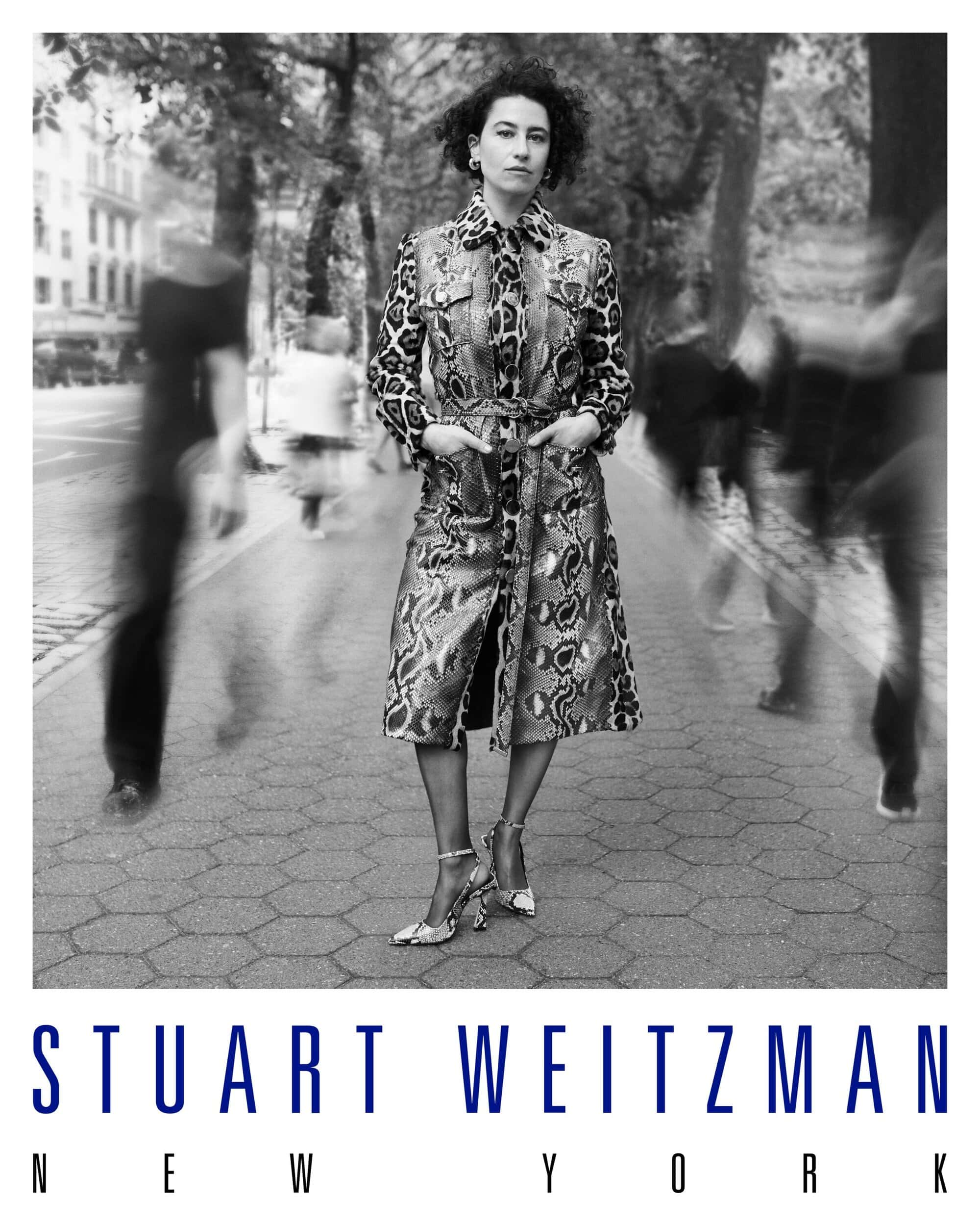 Monochrome portrait of Ilana Glazer standing on a city pavement in a patterned coat with blurred figures passing by.