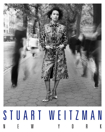 Monochrome portrait of Ilana Glazer standing on a city pavement in a patterned coat with blurred figures passing by.
