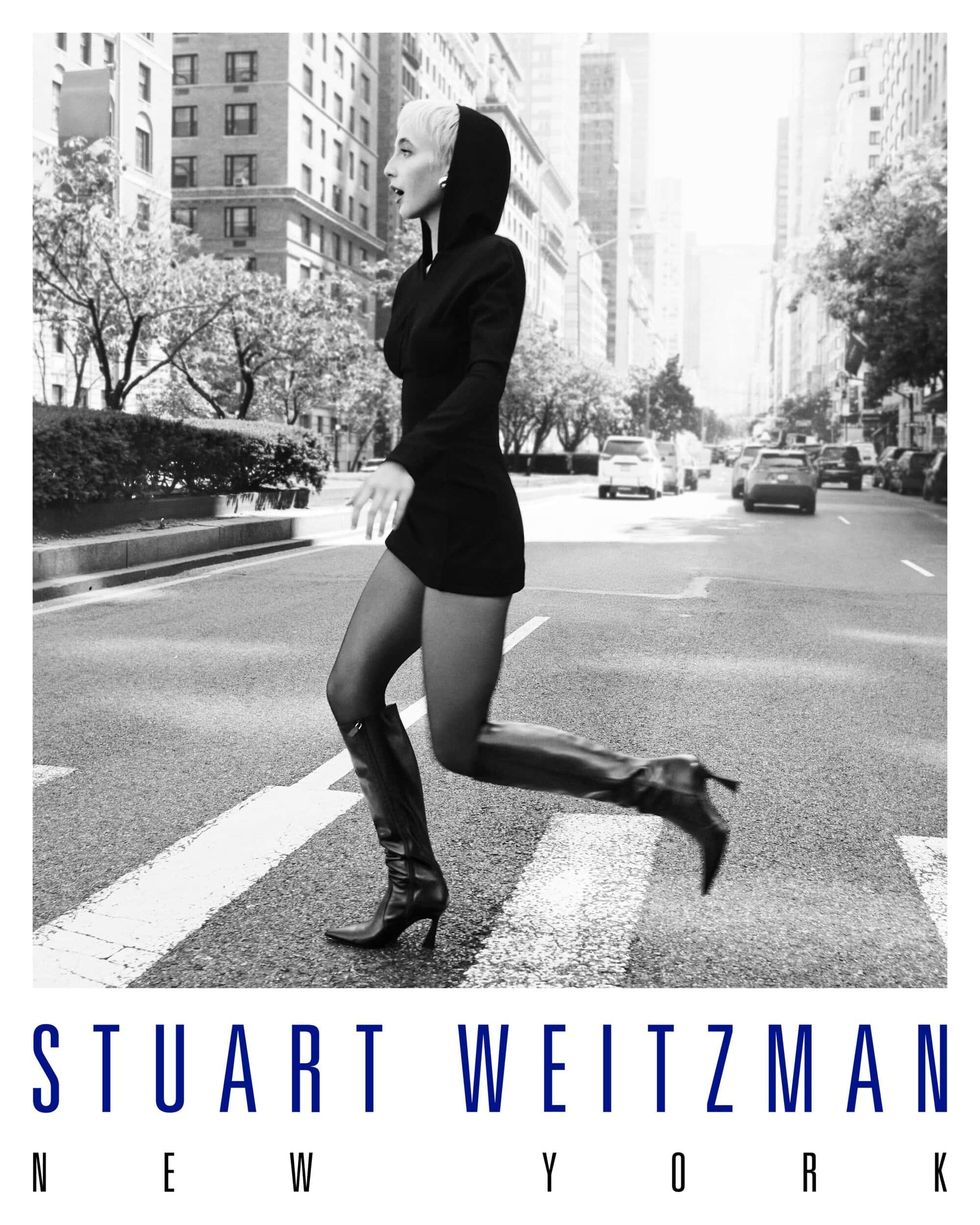 Woman in black hooded dress and tall leather boots walking across a New York City street for Stuart Weitzman.