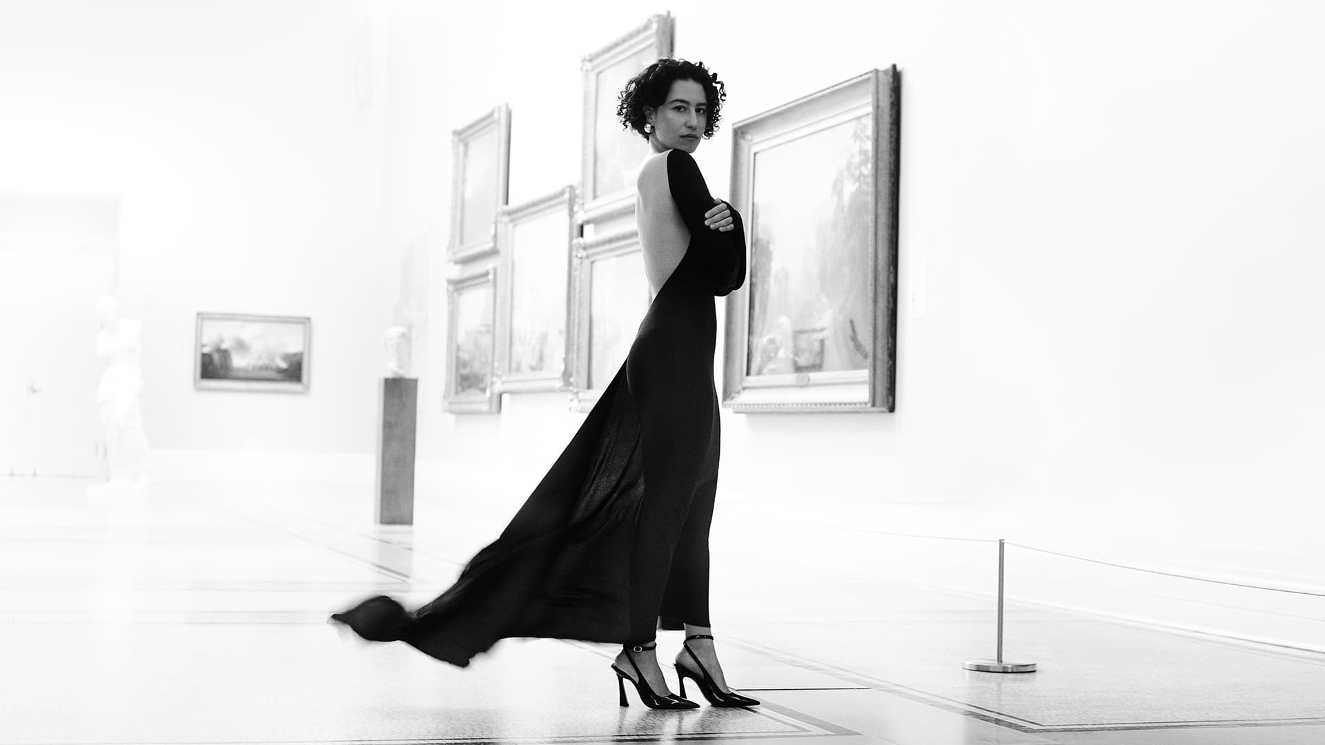 Black and white portrait of a woman in a long black backless dress posing inside a museum gallery