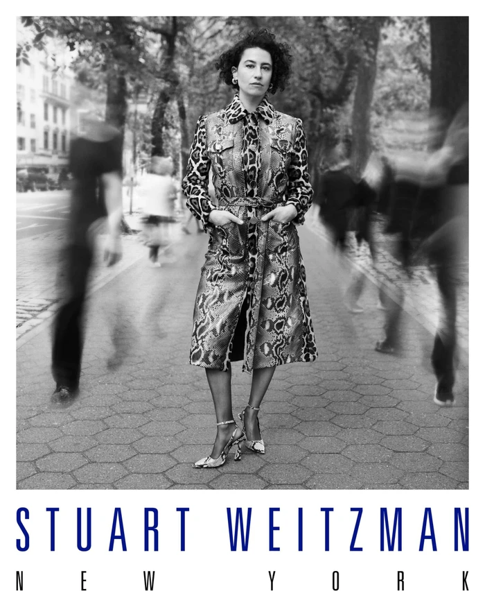 Monochrome portrait of Ilana Glazer standing on a city pavement in a patterned coat with blurred figures passing by.