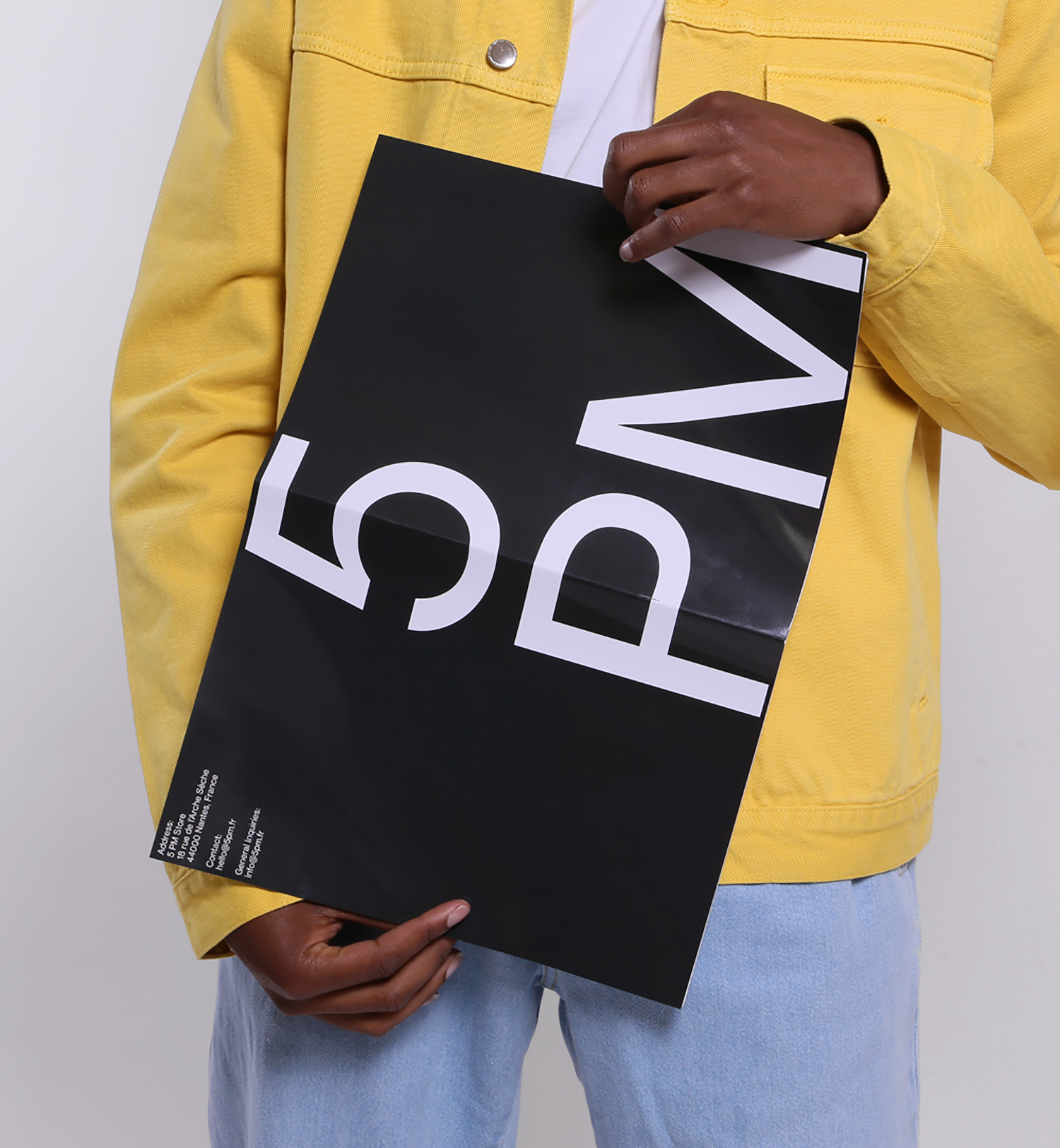Person wearing yellow denim jacket and light blue jeans holding a black poster with bold 5 PM typography