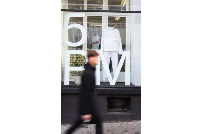 A motion-blurred figure walking past a retail storefront featuring large typography spelling 5PM and a mannequin in white apparel.