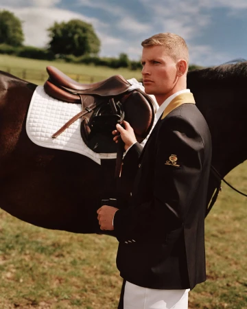Man wearing a black riding jacket standing next to a saddled horse in a grassy field