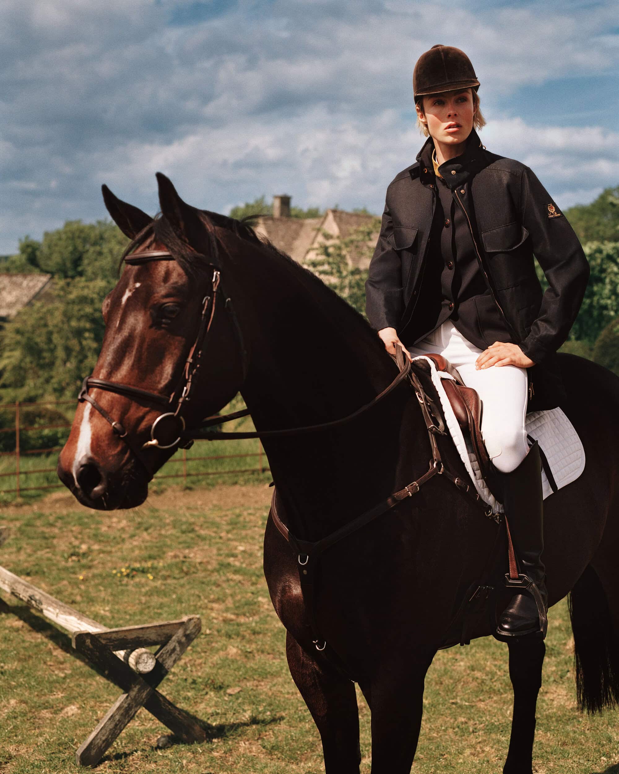 Model Edie Campbell wearing an equestrian jacket and riding helmet while mounted on a dark brown horse outdoors.