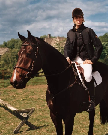 Model Edie Campbell wearing an equestrian jacket and riding helmet while mounted on a dark brown horse outdoors.