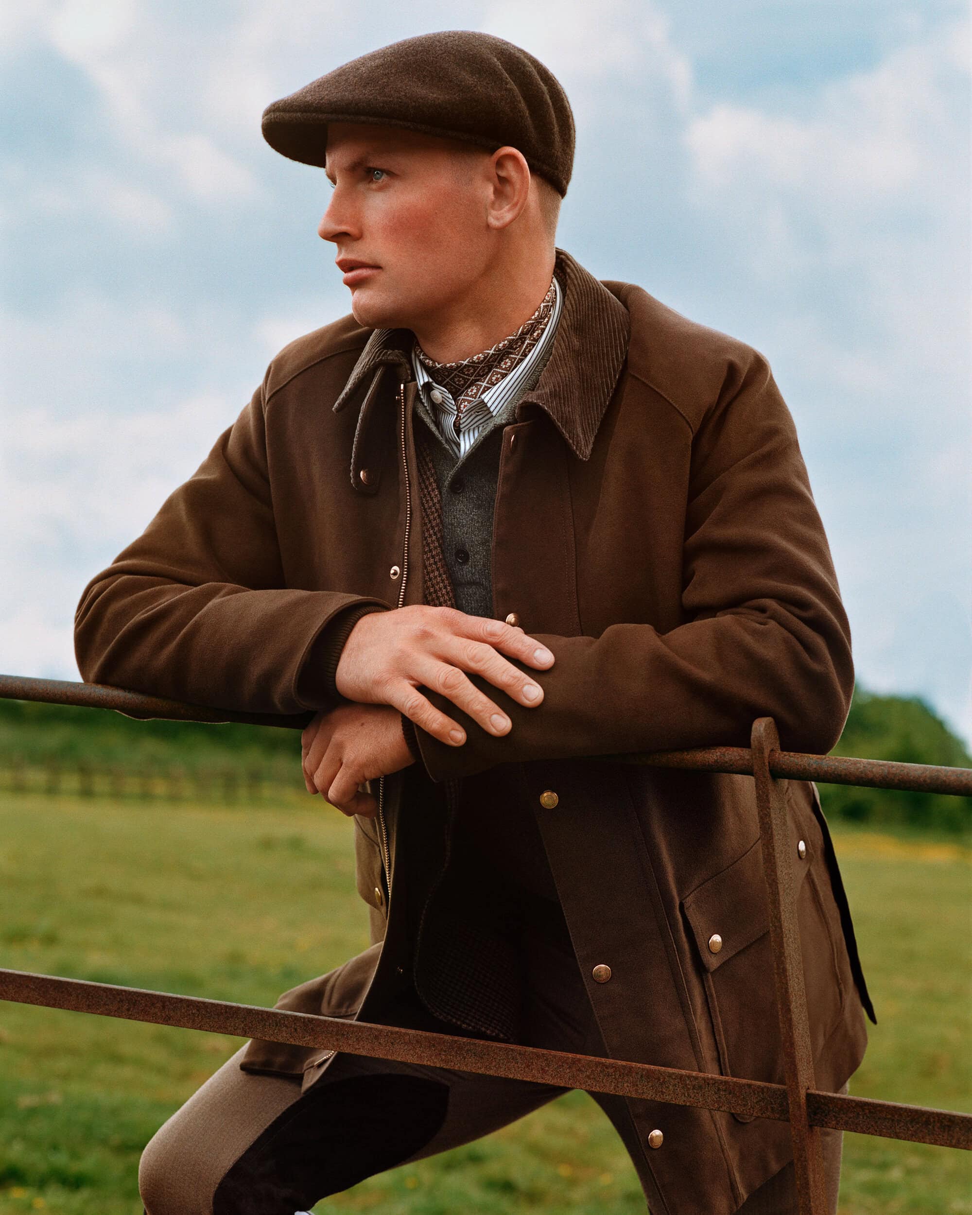 Man wearing a brown jacket and flat cap leaning against a metal fence outdoors in a grassy field under a cloudy sky.