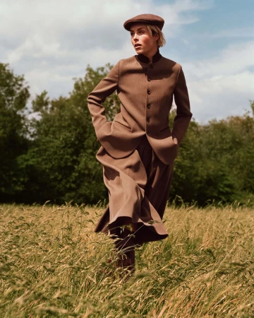 Model wearing brown wool suit and flat cap walking through a tall grass field outdoors
