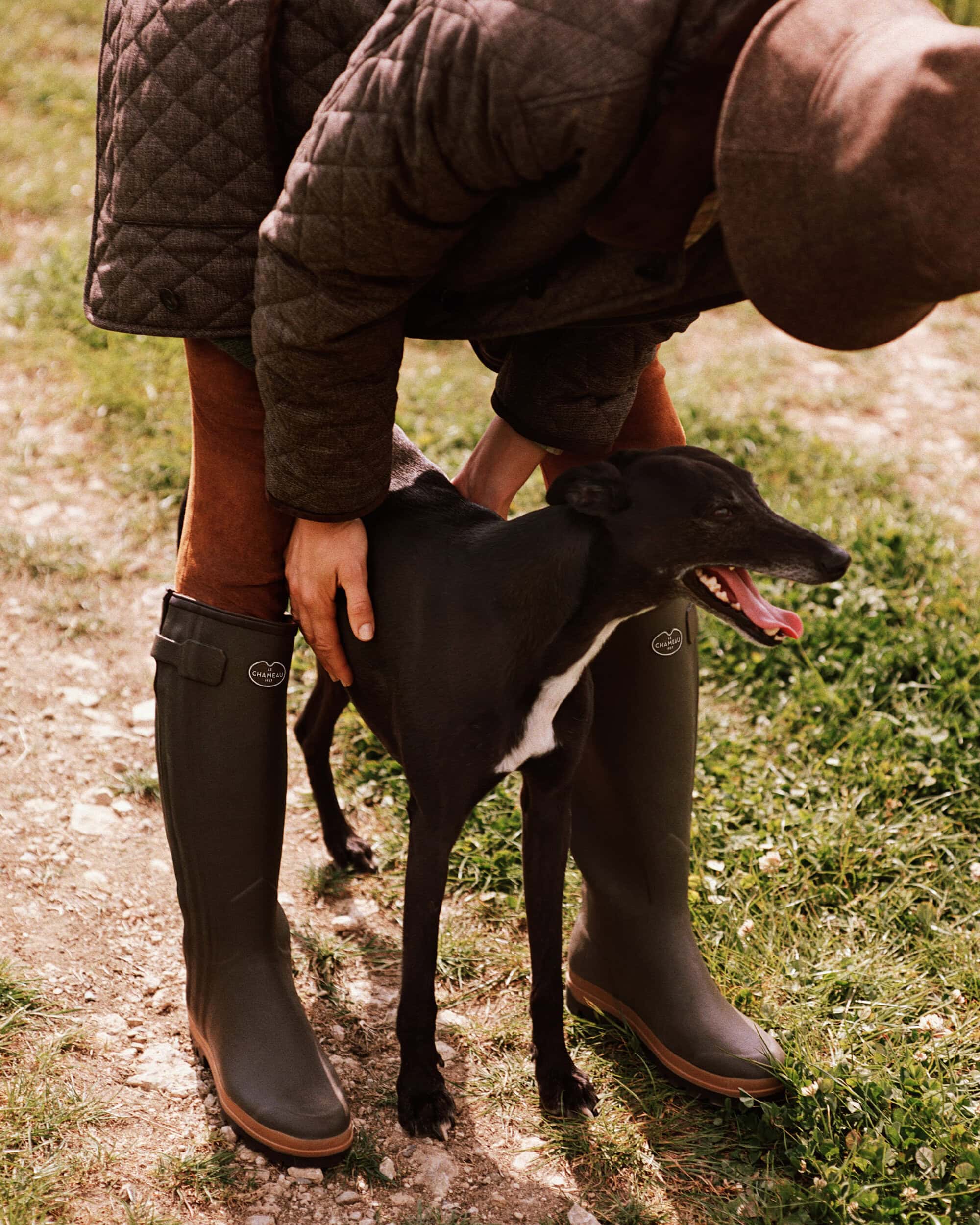 Model wearing a quilted jacket and rubber boots leaning down to pet a black dog on a grassy path.