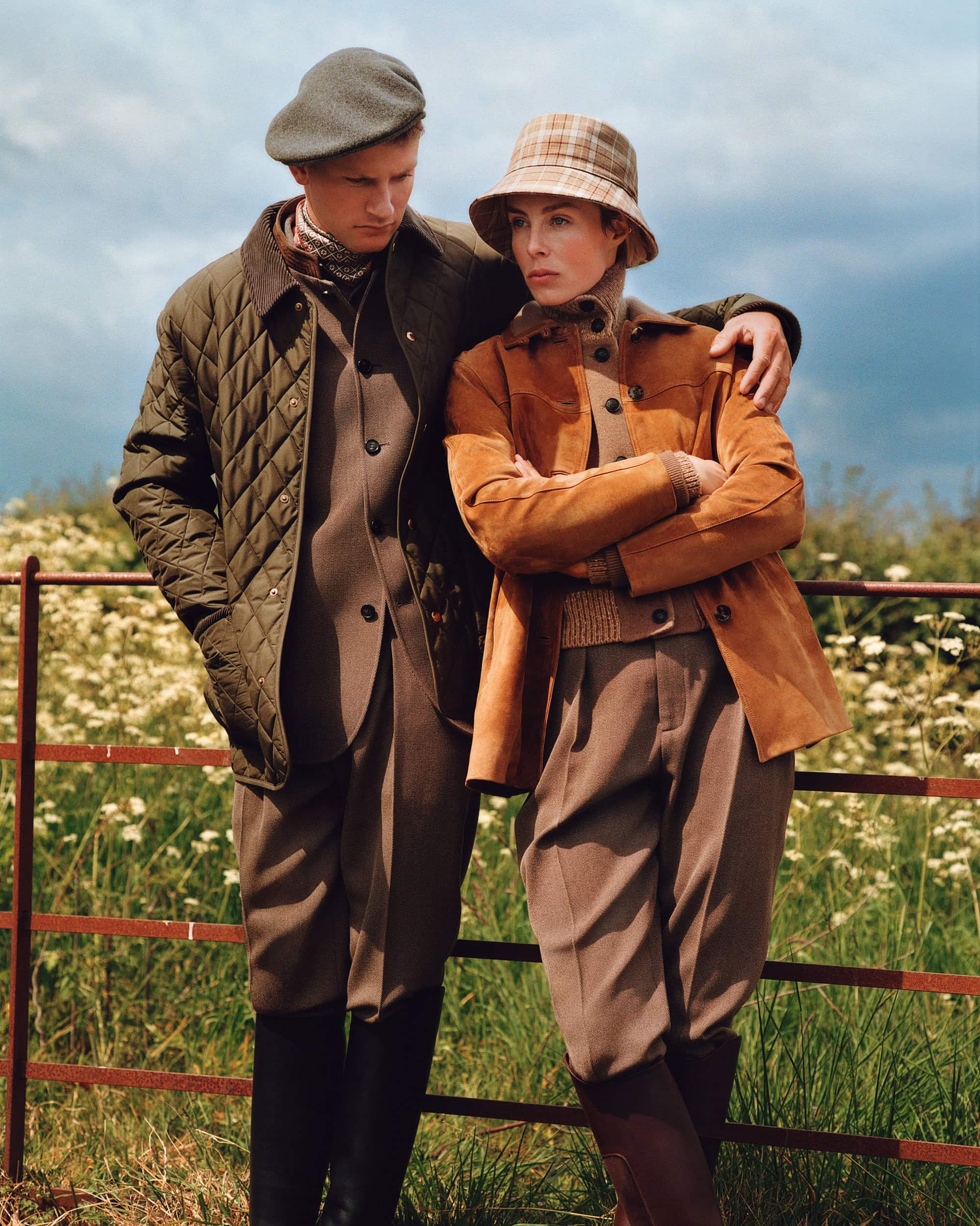 Two models outdoors leaning against a red metal gate wearing outdoor apparel including a quilted jacket and plaid bucket hat.