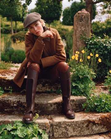 Model Edie Campbell wearing a brown coat, riding boots and cap, seated on stone steps outdoors in an ad campaign by Alasdair McLellan.
