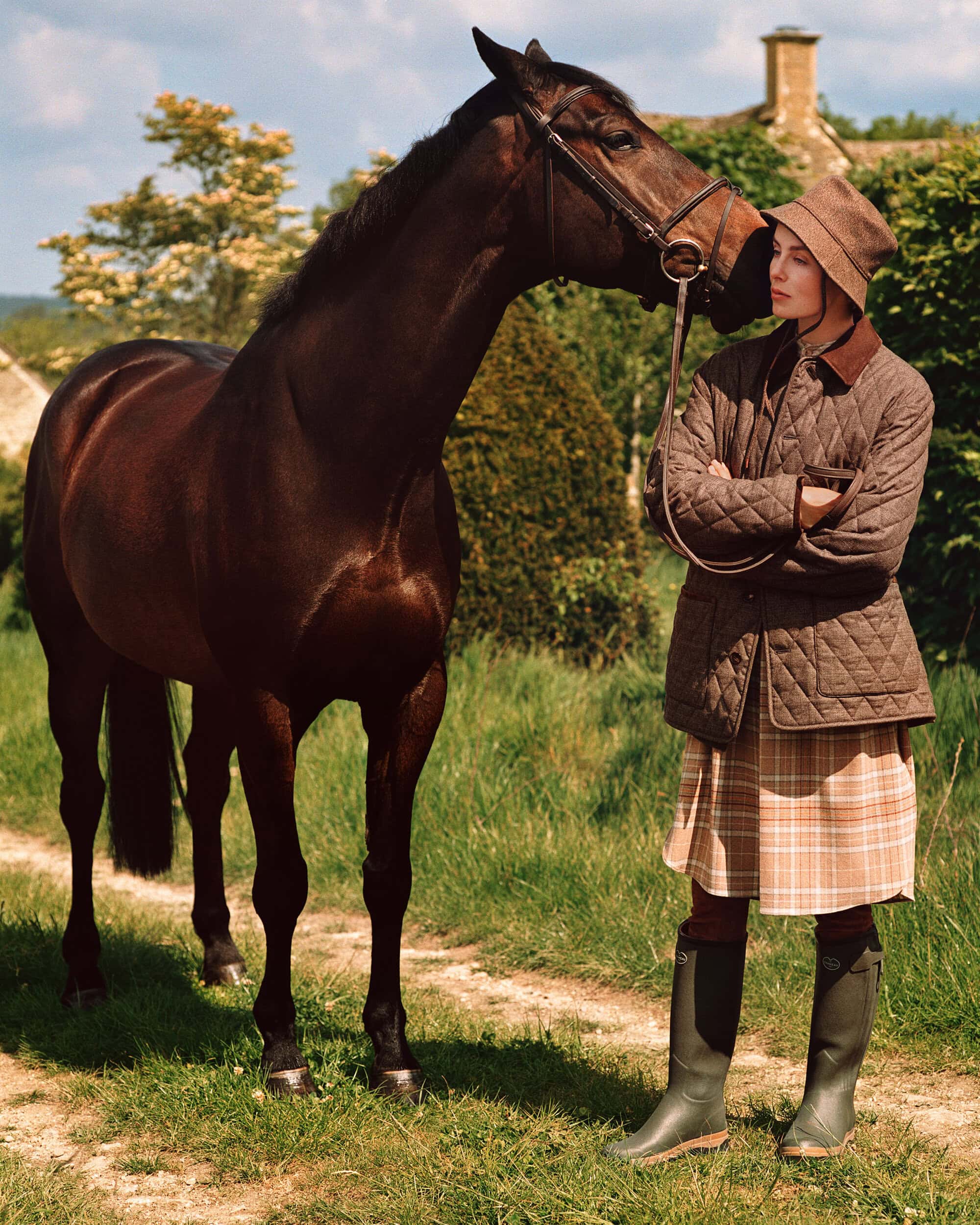 Model standing next to a dark brown horse outdoors, wearing a quilted jacket and tall green boots.