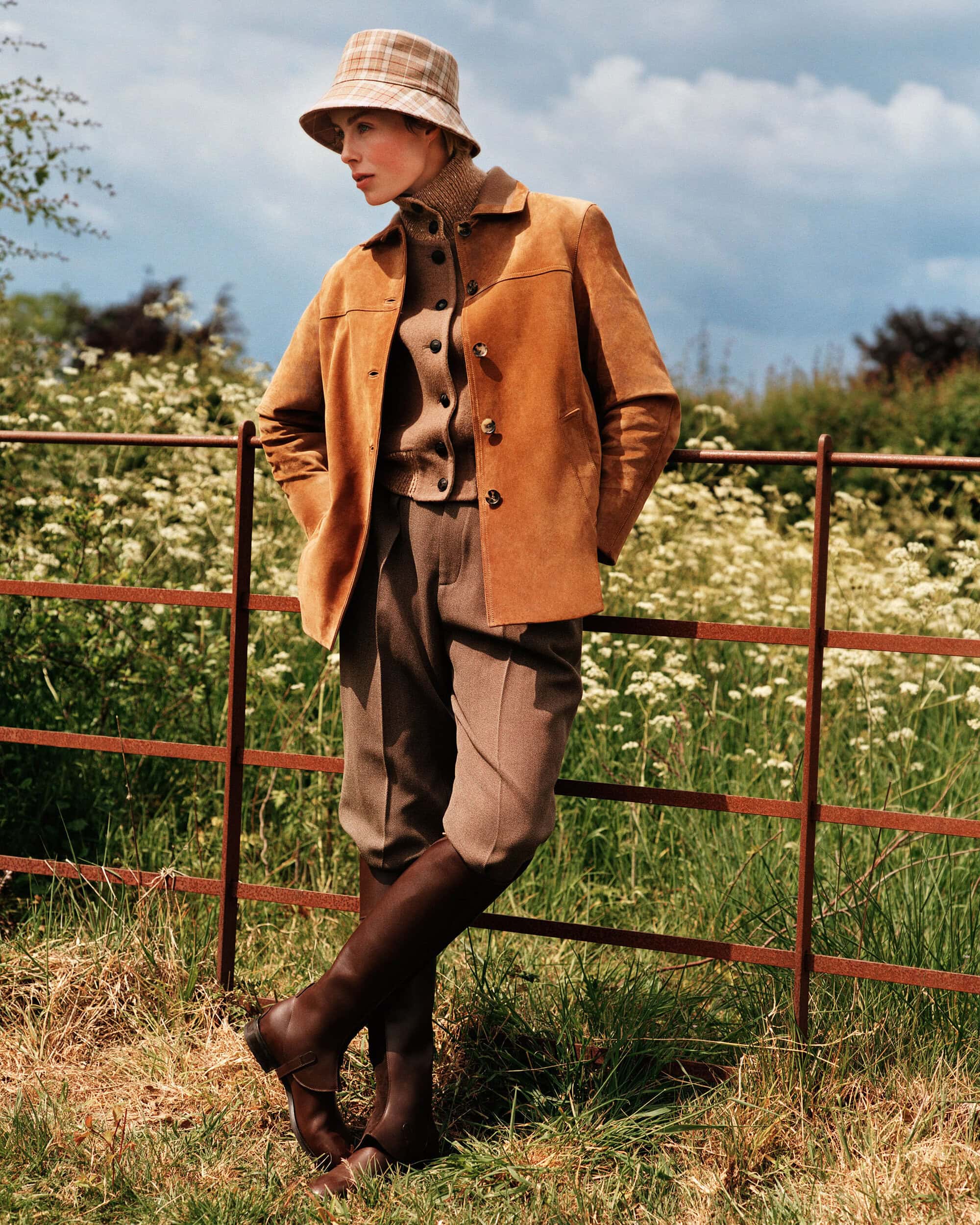 Model Edie Campbell in a brown suede jacket and plaid bucket hat leaning on a fence outdoors for Loro Piana