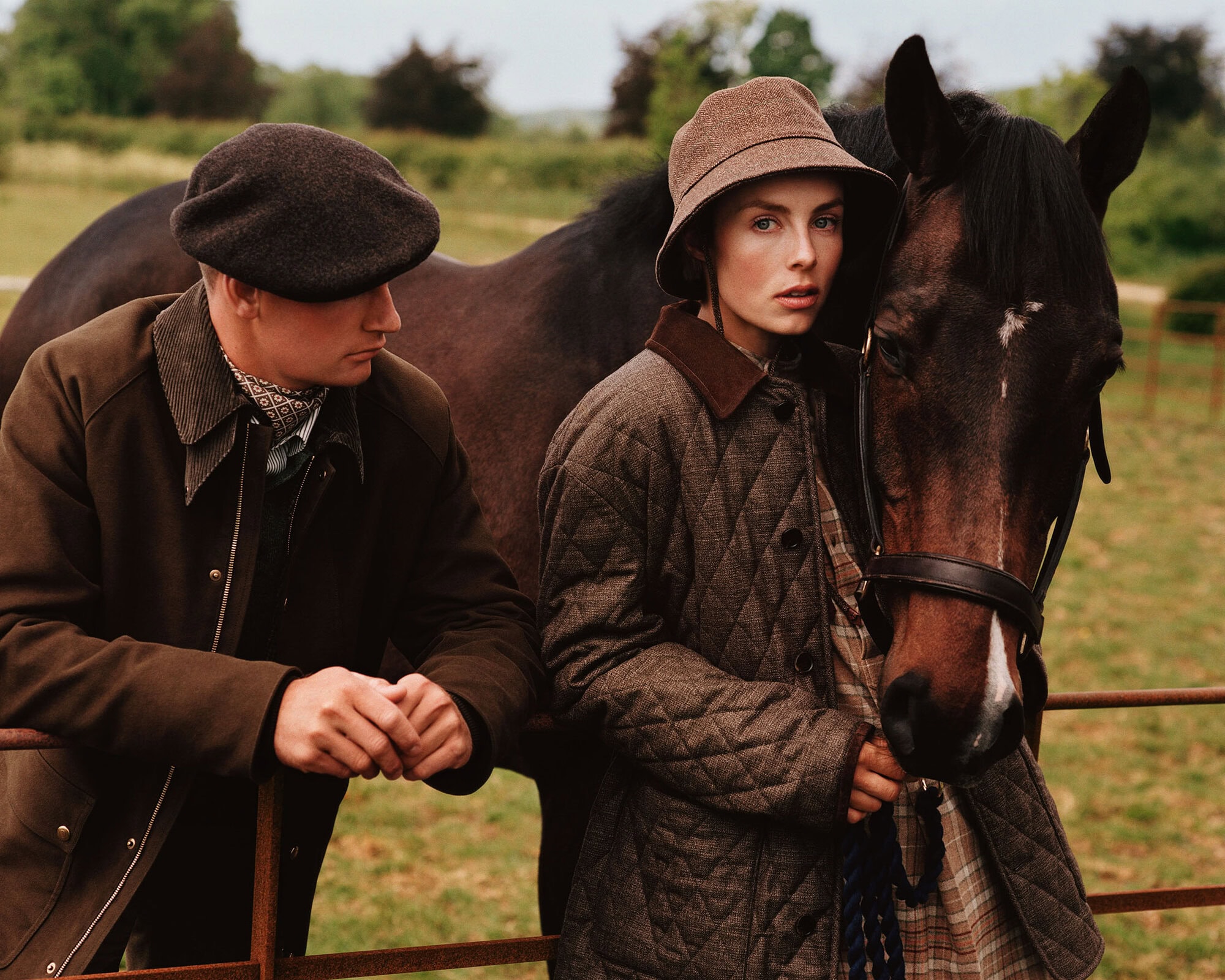 Model in a brown quilted coat and bucket hat standing next to a horse outdoors, with a man in a flat cap leaning on a metal fence.