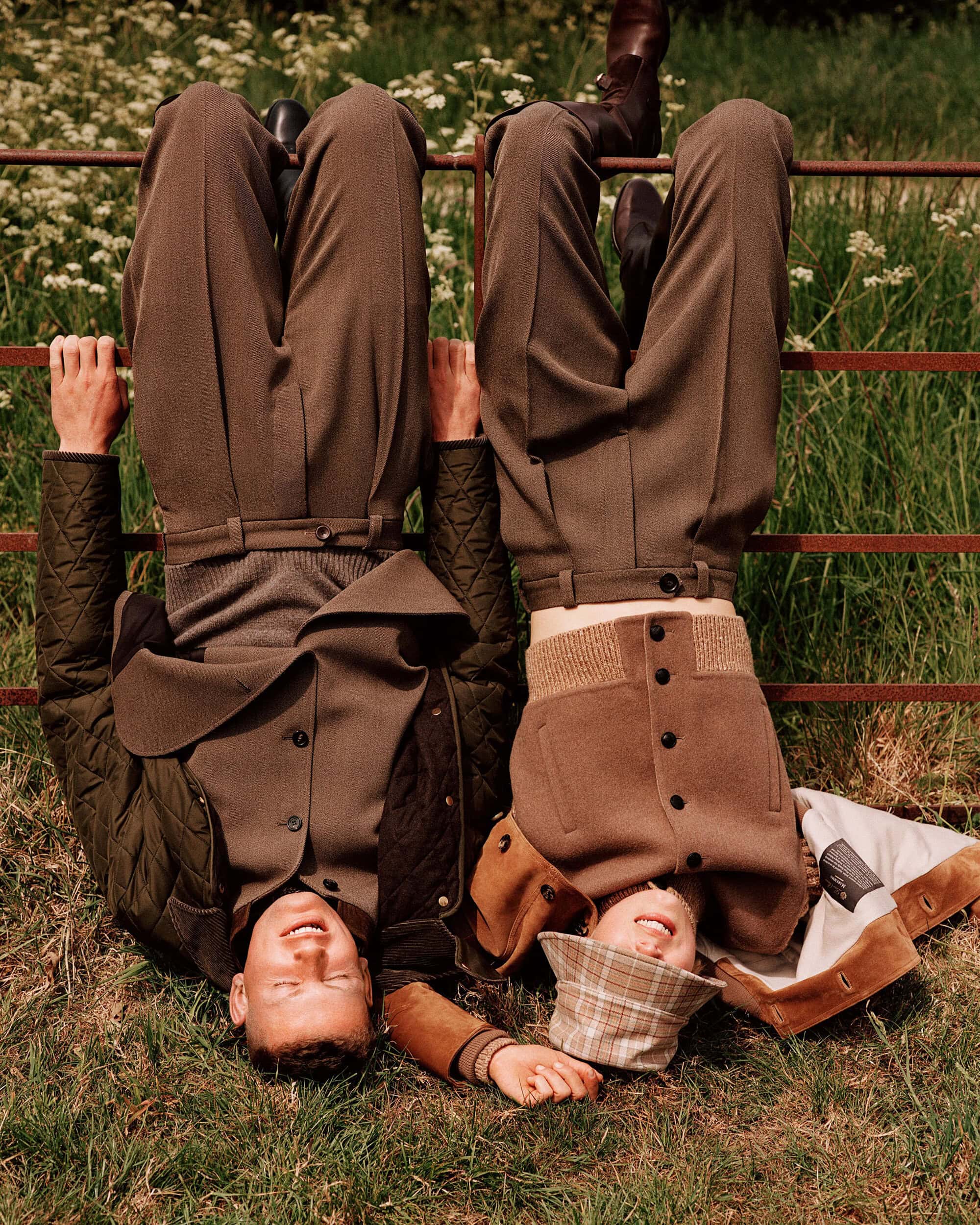 Two models hanging upside down from a metal gate wearing brown trousers and jackets in a grassy field.