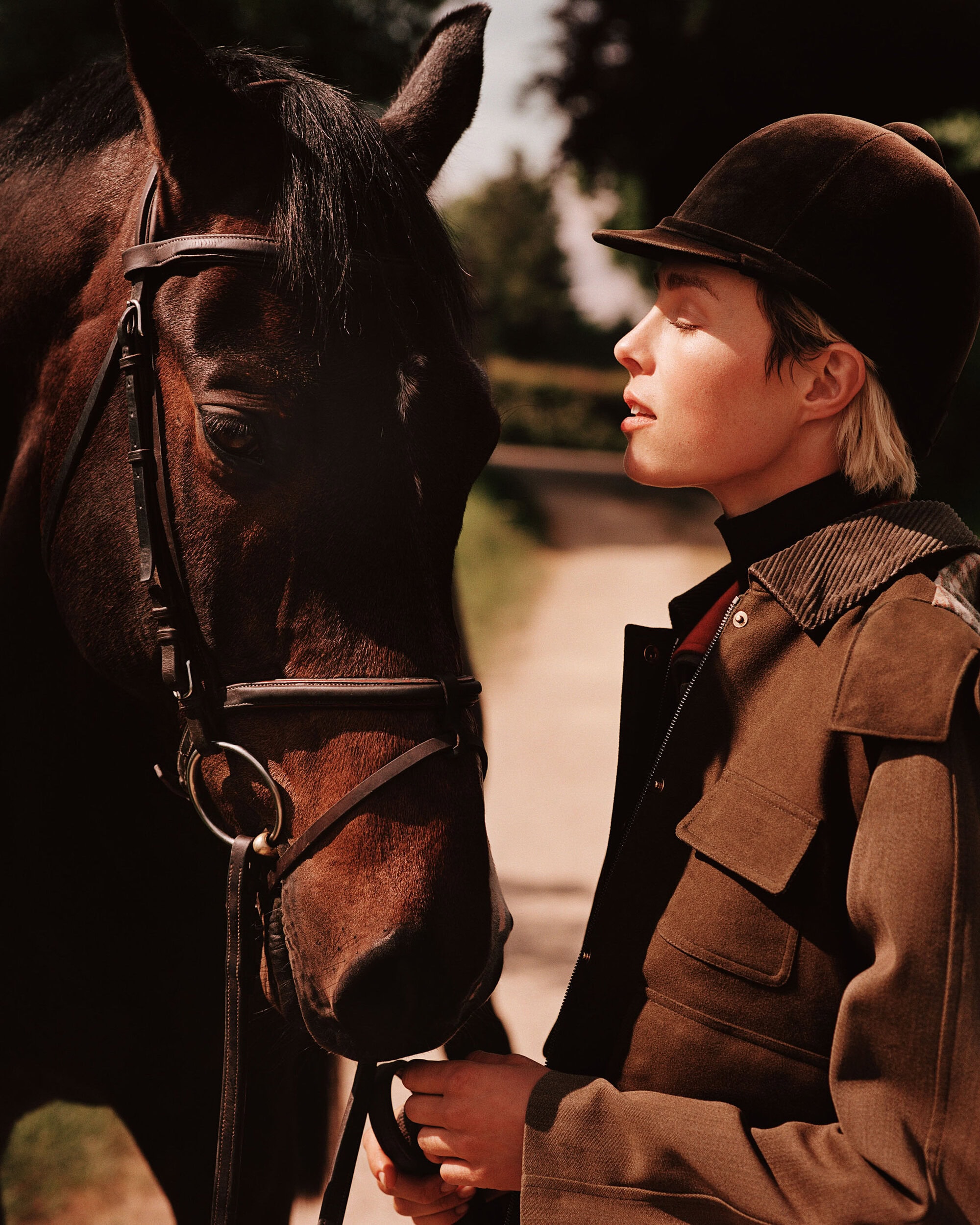 Model Edie Campbell wearing an equestrian jacket and helmet standing beside a horse outdoors.