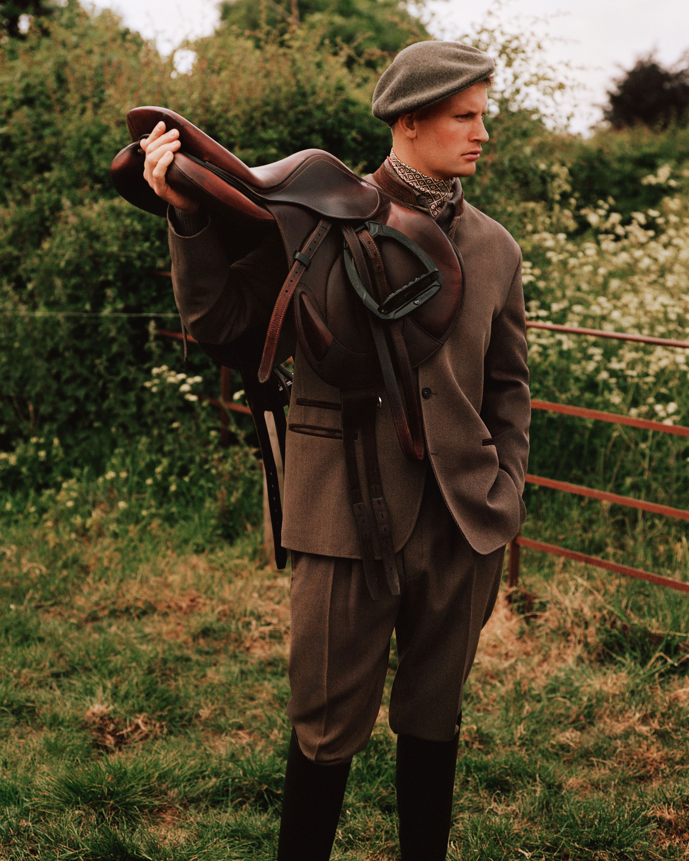 Man wearing a brown tweed suit and cap holding a leather saddle outdoors in a rural setting.