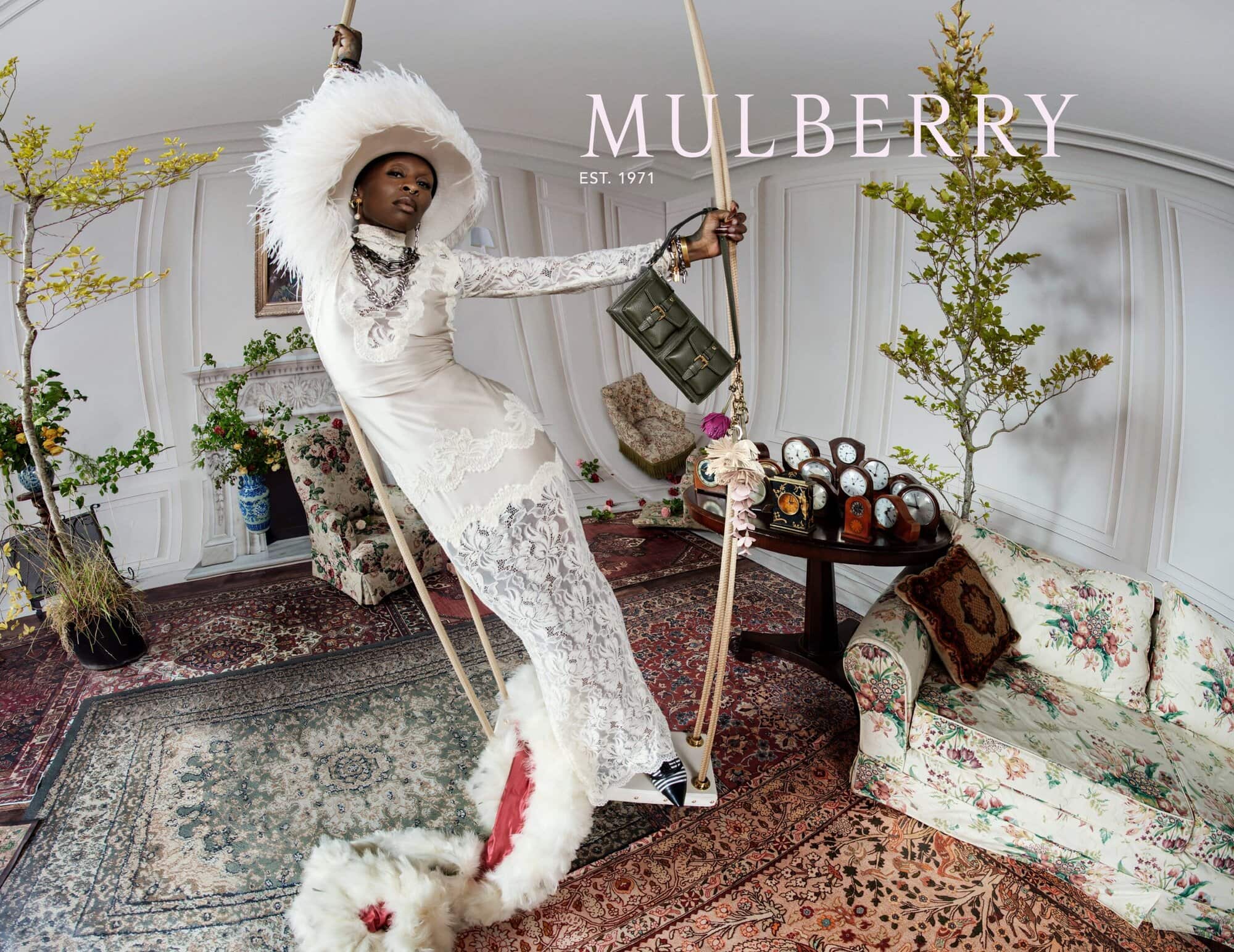 Cynthia Erivo wearing a white lace dress and feathered hat on a swing in a surreal drawing room for Mulberry