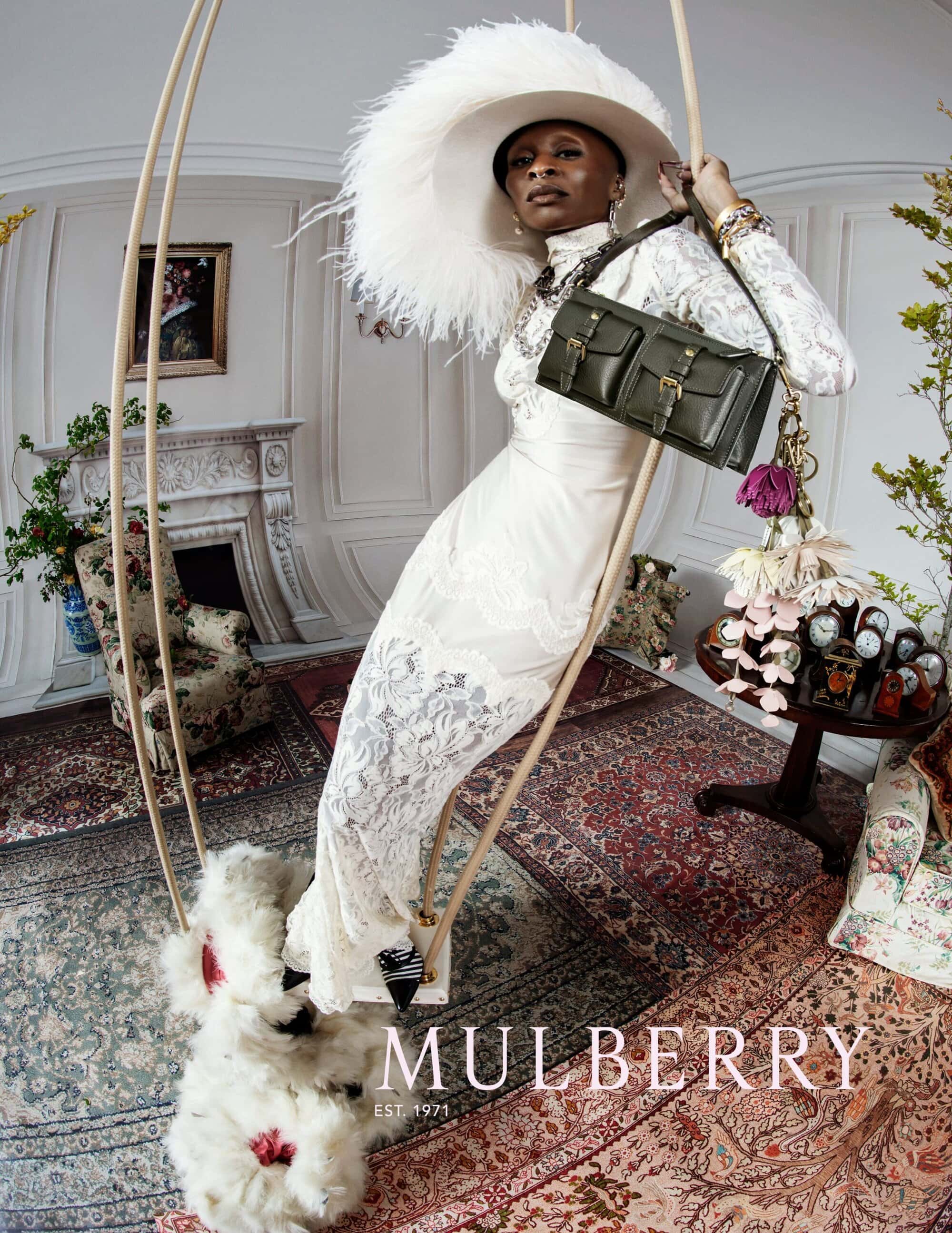 Cynthia Erivo wearing a wide feather-trimmed hat and white lace dress, holding a green shoulder bag while posing on an indoor swing