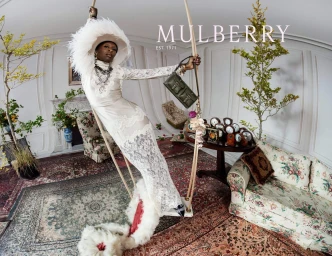Cynthia Erivo wearing a white lace dress and feathered hat on a swing in a surreal drawing room for Mulberry