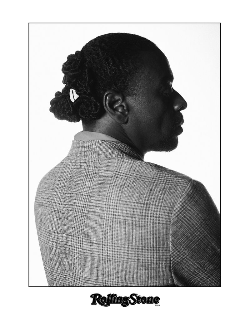 Portrait of an adult male with dark hair in locs, wearing a grey and black plaid jacket, back to the camera, on a white background.