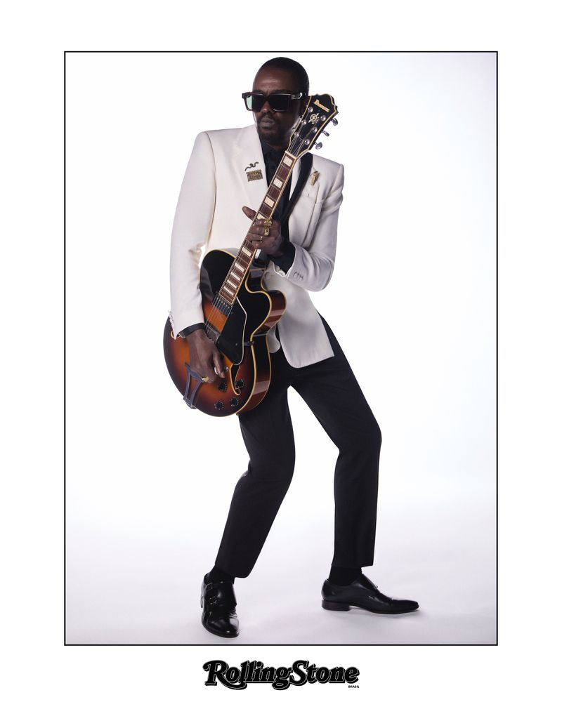 Man in a white suit plays an electric guitar in a studio setting, standing against a white backdrop with a confident stance and sunglasses.