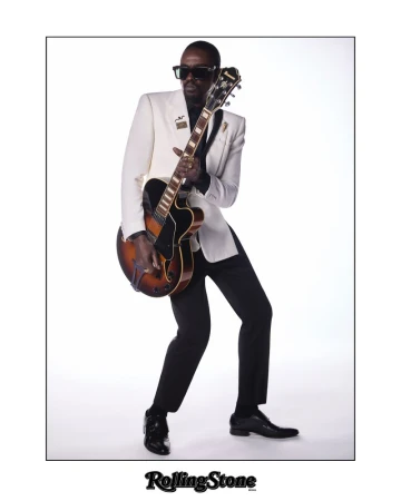 Man in a white suit plays an electric guitar in a studio setting, standing against a white backdrop with a confident stance and sunglasses.