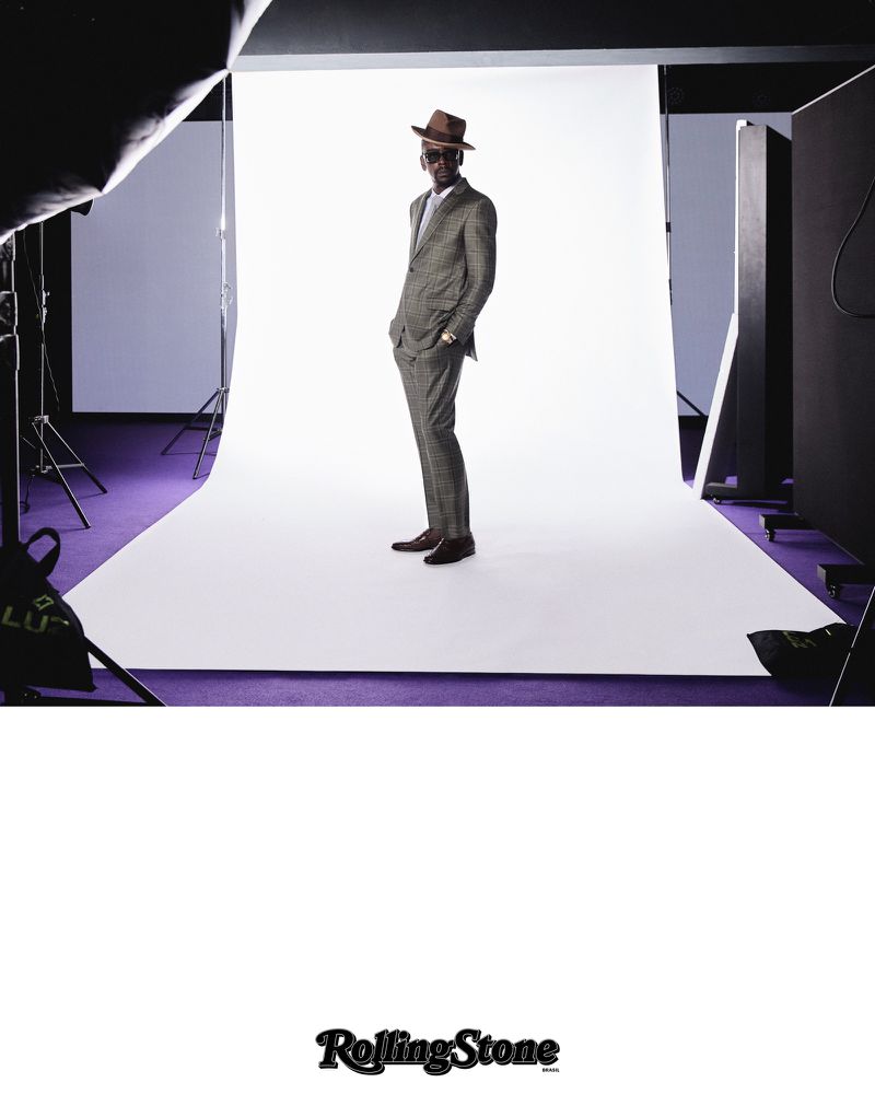 Full-length shot of a stylish adult man in a checkered suit and hat, standing in a photo studio with white backdrop and purple floor.