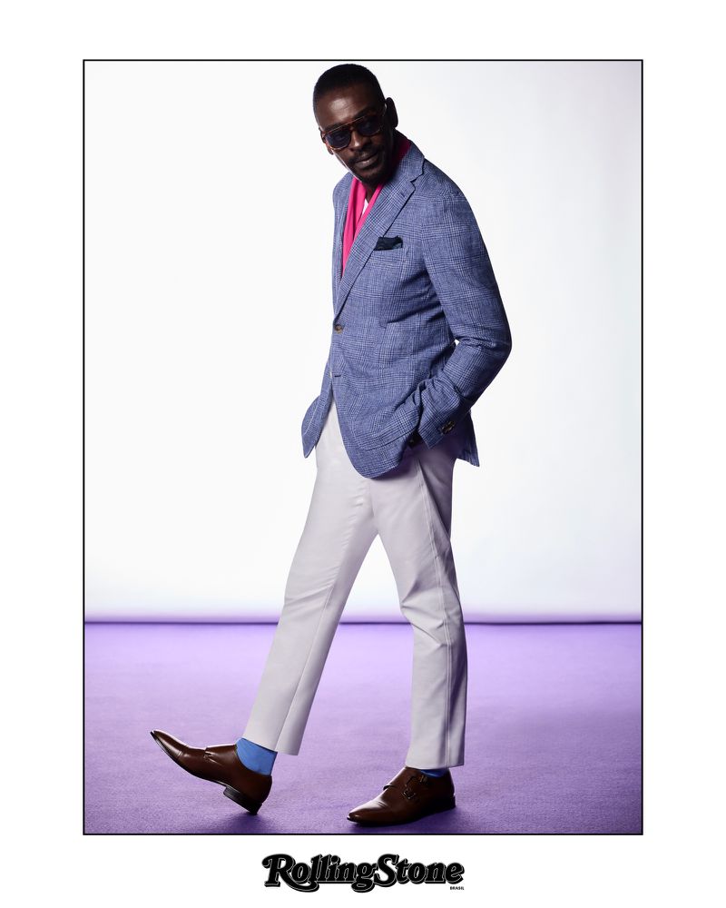 Full shot of a Black male fashion model in a blue jacket, white pants, sunglasses, and brown shoes posing on a graded white to purple backdrop.