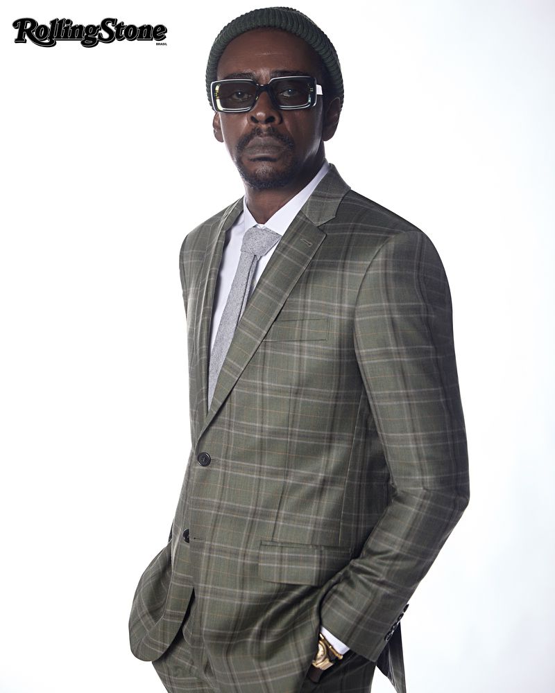 Portrait of a stylish adult man in a plaid suit, olive beanie, and sunglasses, posing against a white background.