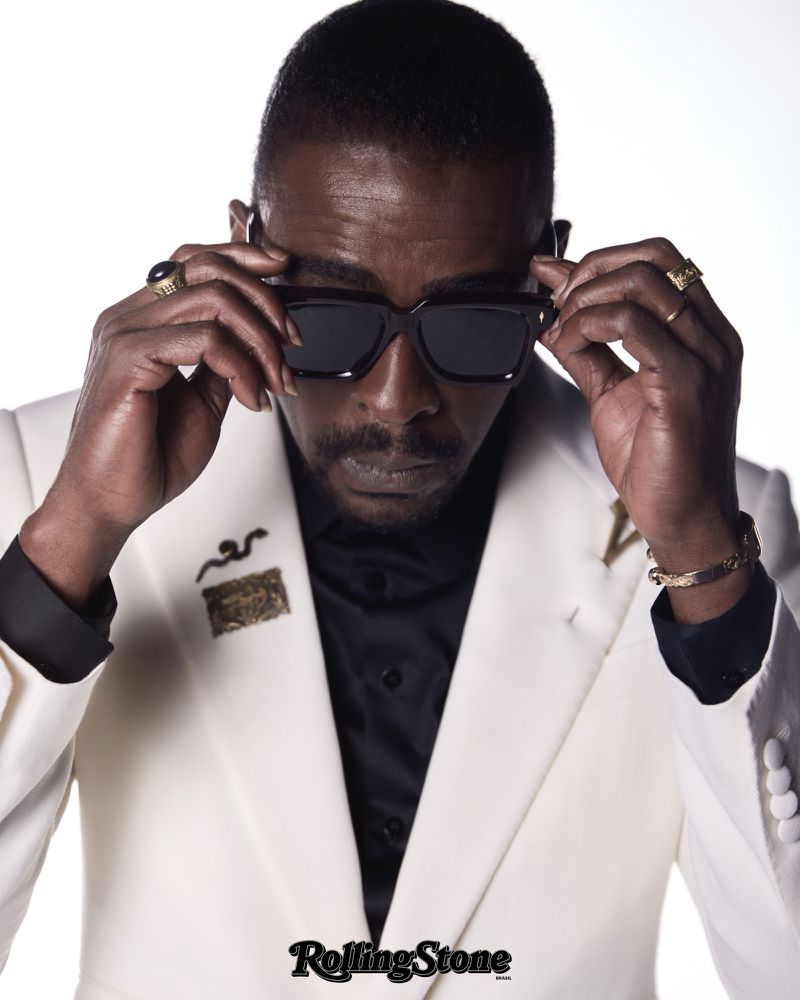 Portrait of a middle-aged Black male with dark hair wearing sunglasses, a black shirt, white suit and jewelry with a white background.