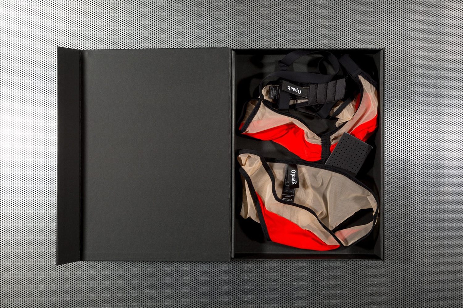 Black presentation box containing red and beige undergarments with black straps, placed on a textured metal surface.