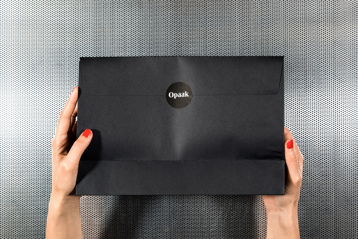 Two hands holding a large black envelope package with a circular sticker against a perforated metal background.