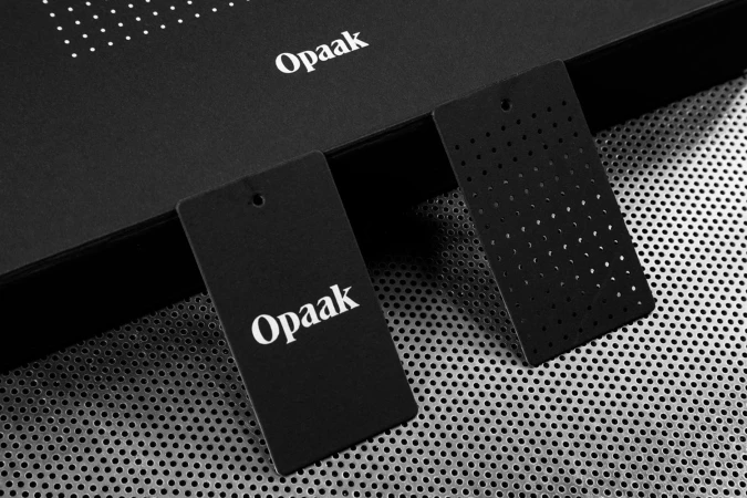 Close-up of black brand tags with white typographic logo and perforated patterns resting on a metallic mesh surface.