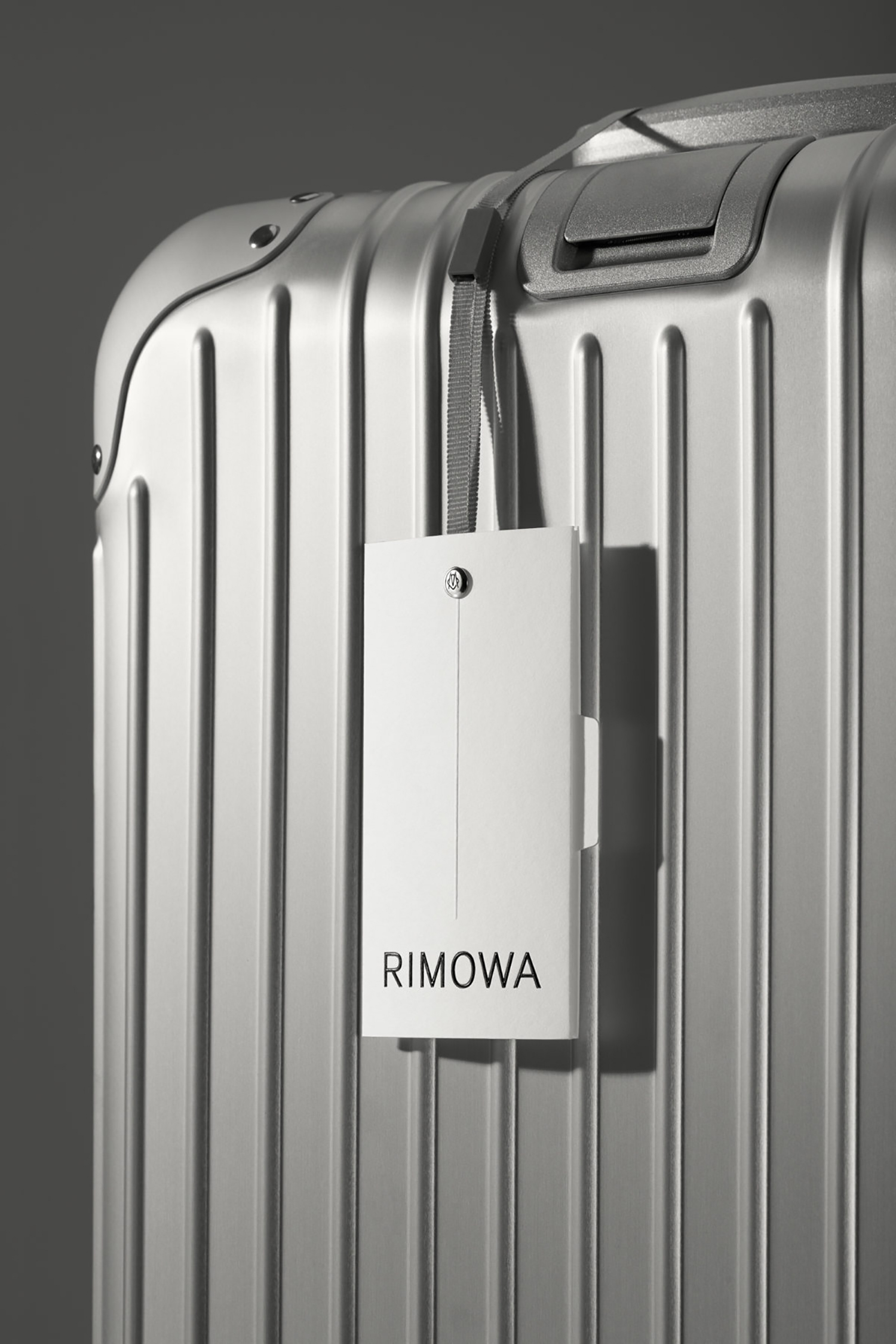 Close-up of a silver grooved suitcase with a white Rimowa luggage tag hanging from the handle against a grey background