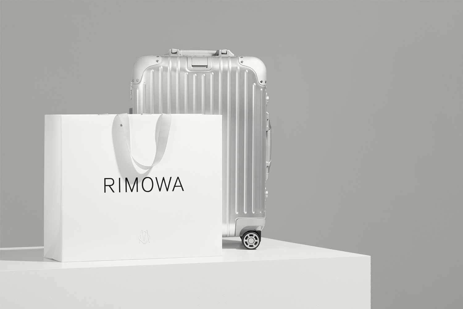 RIMOWA silver aluminum suitcase and white shopping bag with black logo on a white surface against a grey backdrop