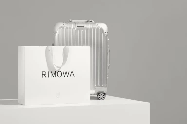RIMOWA silver aluminum suitcase and white shopping bag with black logo on a white surface against a grey backdrop