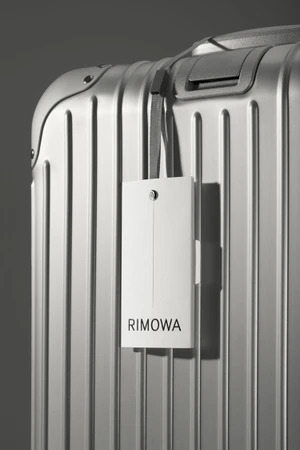 Close-up of a silver grooved suitcase with a white Rimowa luggage tag hanging from the handle against a grey background