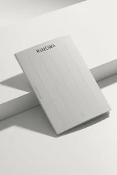 RIMOWA branded manual booklet resting on angled white stepped platform surfaces under soft studio lighting.