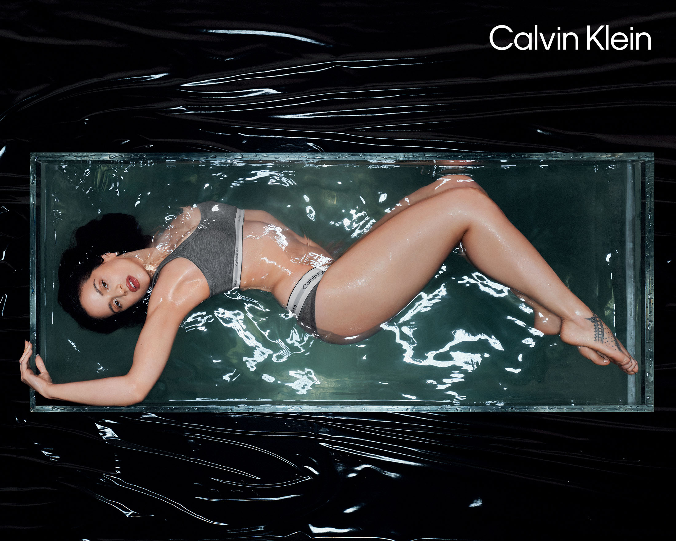 Rosalía lying submerged in a glass water tank wearing grey underwear for a Calvin Klein underwear advertising campaign.
