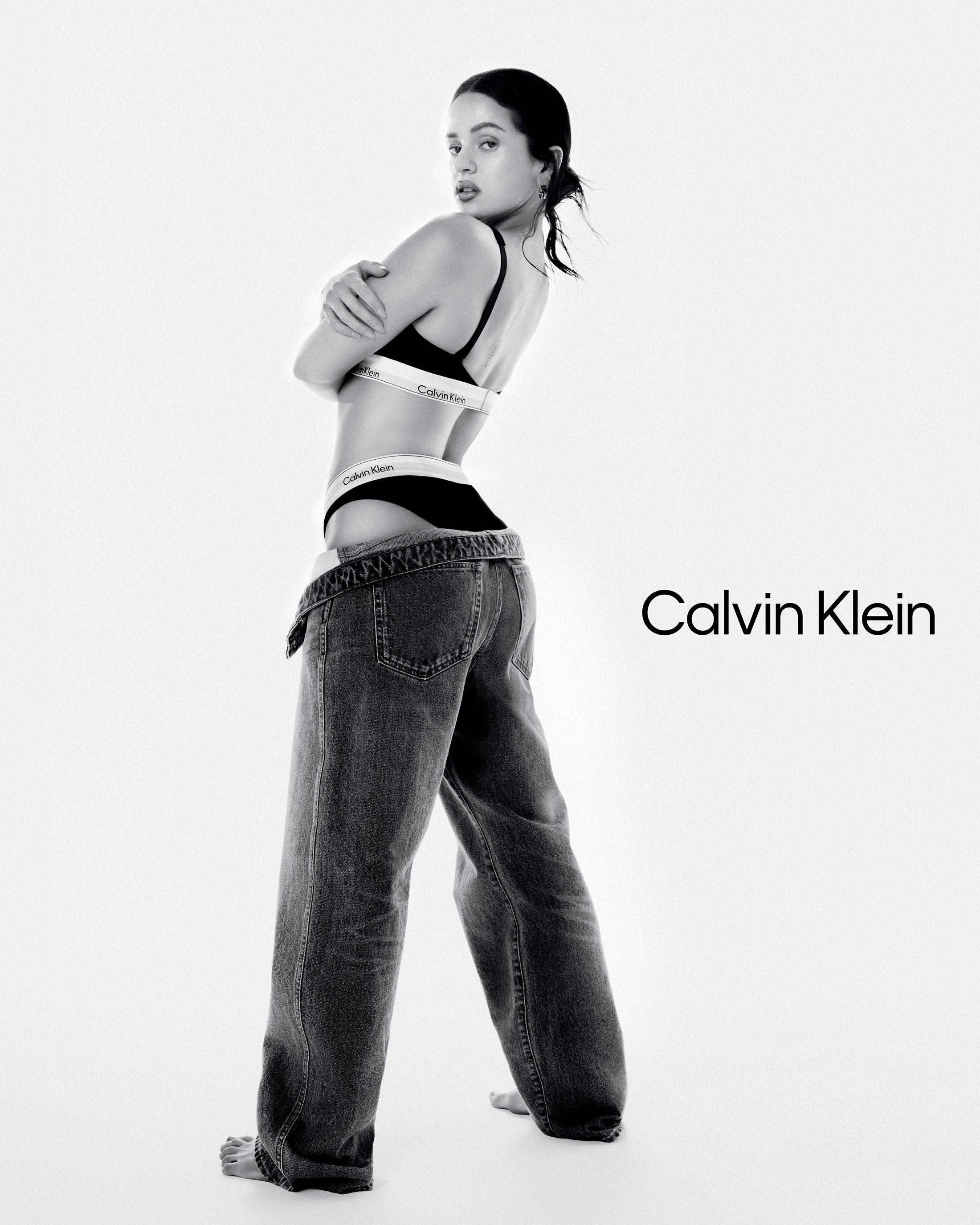 Rosalía posing in Calvin Klein black underwear and baggy denim jeans against a white background