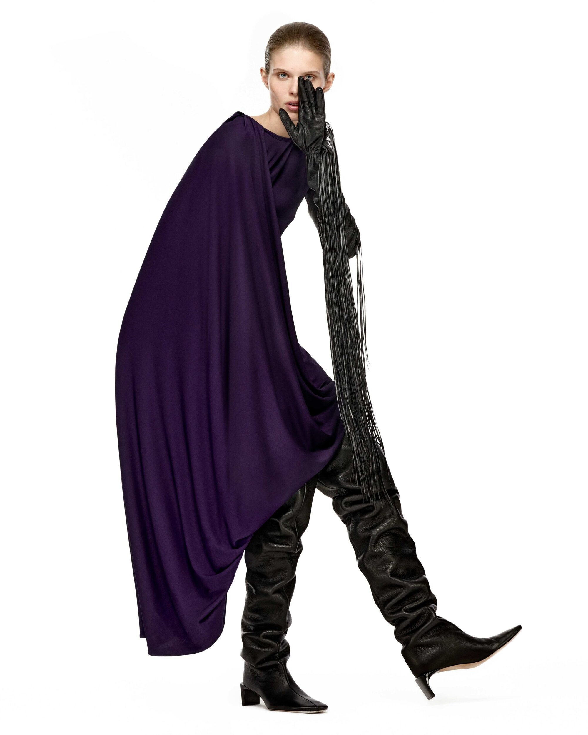 Model in a deep purple draped dress and tall black boots holding a gloved hand with fringe over her face