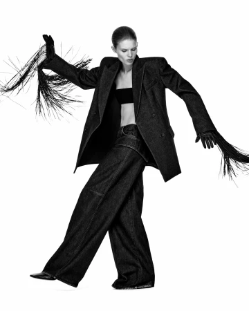 Model wearing a structured dark blazer with fringed sleeves and matching wide-legged trousers against a plain white backdrop.