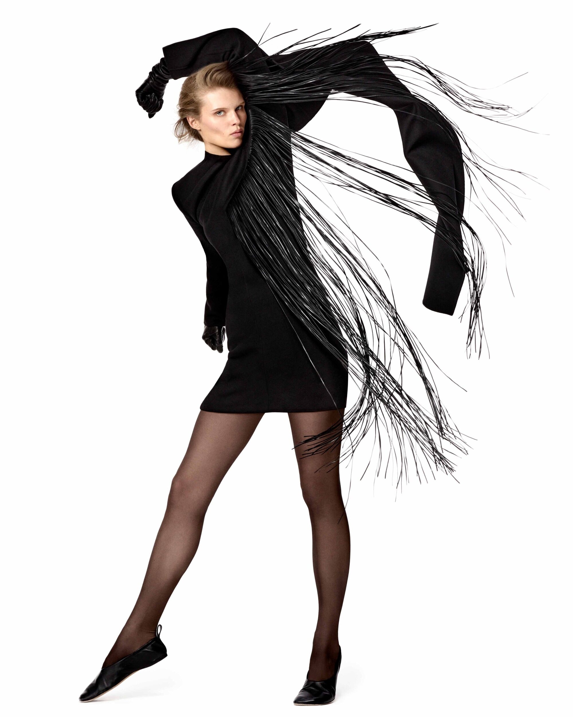 Model in black dress with dramatic fringe sleeves and dark tights posing against white background