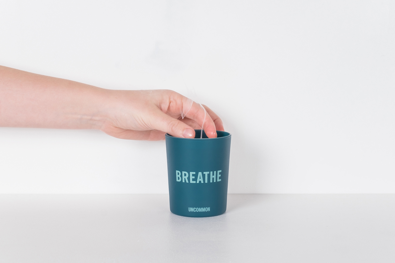 A hand pulling a transparent tag from a teal ceramic cup bearing the word BREATHE against a plain white background.