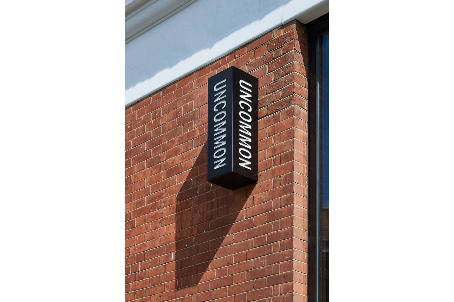 Black rectangular sign with white Uncommon typography mounted on a red brick building wall