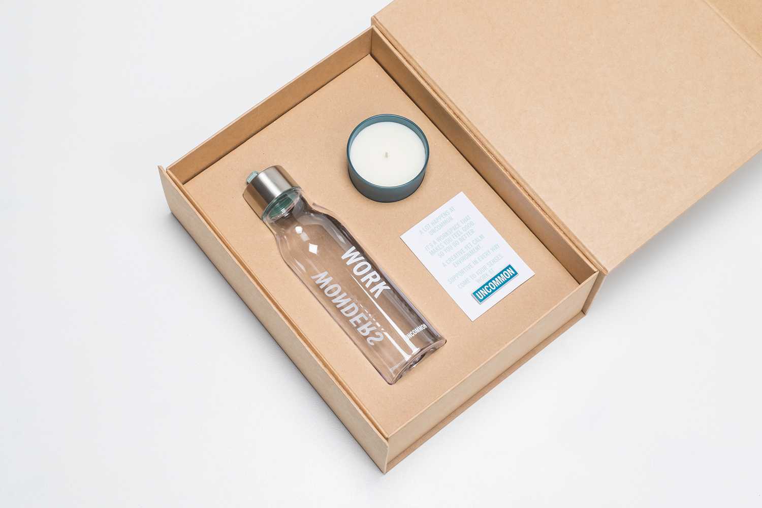 An open cardboard gift box containing a glass water bottle, a round candle, and a branded card for Uncommon.
