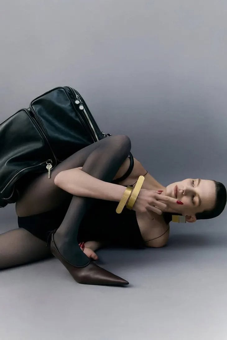 Model lying on grey studio floor wearing black bodysuit and dark tights next to a large black leather bag